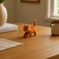 Small orange cat figurine on a wooden table with a mug, books, and plant in the background.
